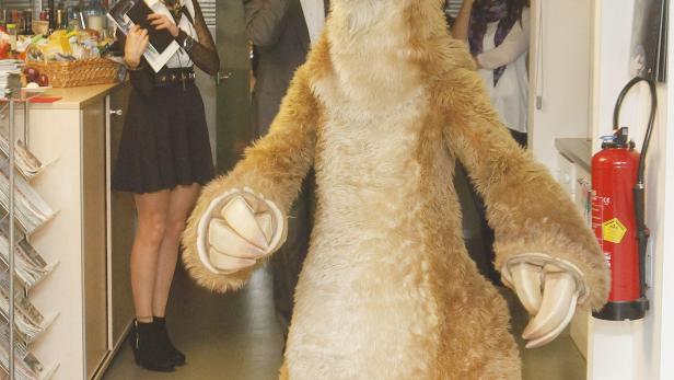 Ein Sid-Faultier-Kostüm steht in einem Büro, während Leute im Hintergrund zusehen.