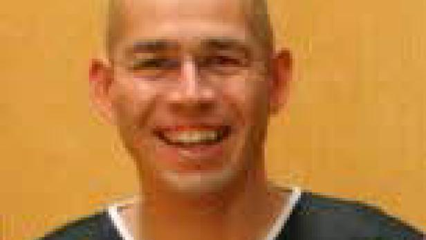 Ein lächelnder, glatzköpfiger Mann mit Brille in einem Sporttrikot.