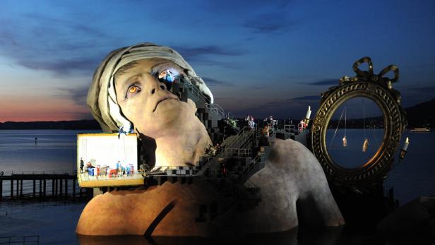 Eine riesige Frauenkopf-Skulptur dient als Bühne auf dem Wasser, mit Schauspielern und Requisiten darauf.