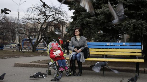 Eine Frau sitzt mit einem Kind im Kinderwagen auf einer Bank, umgeben von Tauben.