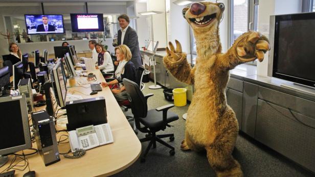 Ein Sid-Faultierkostüm steht in einem Büro mit mehreren Mitarbeitern.