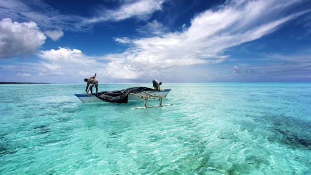 Zwei Fischer arbeiten auf einem Auslegerboot im türkisfarbenen Wasser.