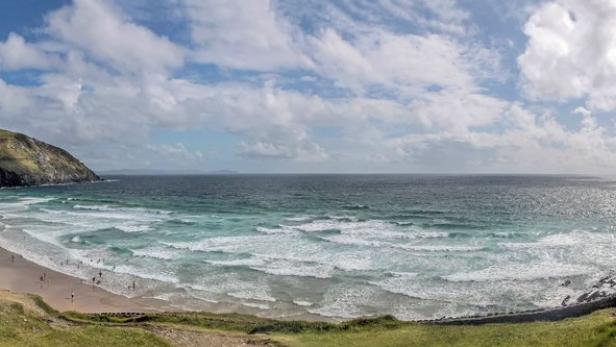Panoramaaufnahme eines Strandes mit grünen Hügeln und türkisfarbenem Wasser.