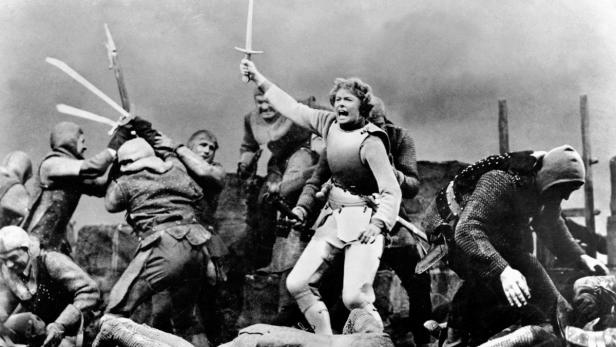 Eine Frau in Rüstung steht inmitten einer Schlacht und hält ein Schwert in die Höhe.