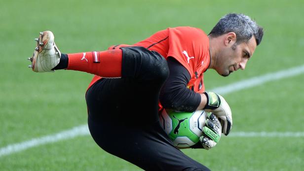 Ein Torwart in rotem Trikot fängt einen Ball auf einem grünen Rasen.
