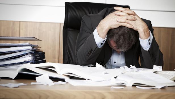Ein gestresster Mann sitzt mit dem Kopf in den Händen an einem Schreibtisch voller Papier.
