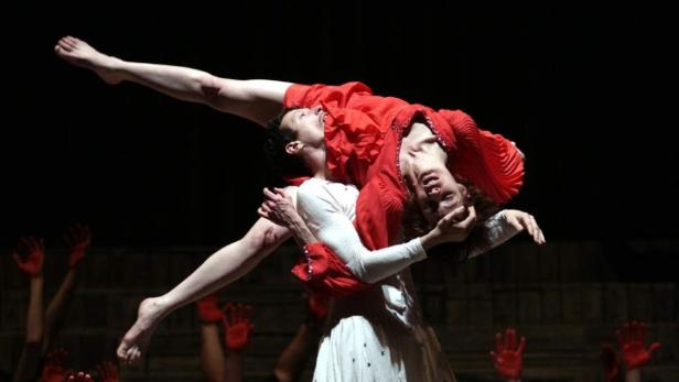 Ein Tänzer hält eine Tänzerin in rotem Kleid über seinem Kopf, während andere Tänzer rote Hände in die Luft strecken.