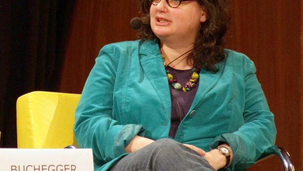 Eine Frau, möglicherweise Buchegger, sitzt auf einem gelben Stuhl und spricht.