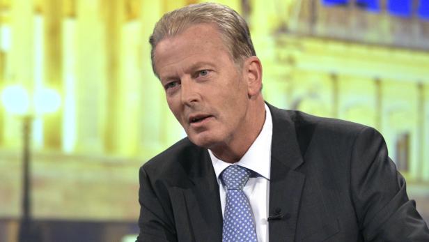 Ein Mann mit Anzug und Krawatte spricht vor einem unscharfen Hintergrund.