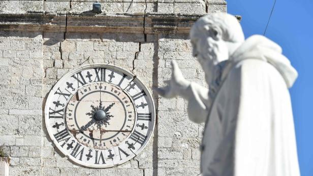 Eine Uhr mit römischen Ziffern an einer steinernen Gebäudefassade, neben einer Statue.