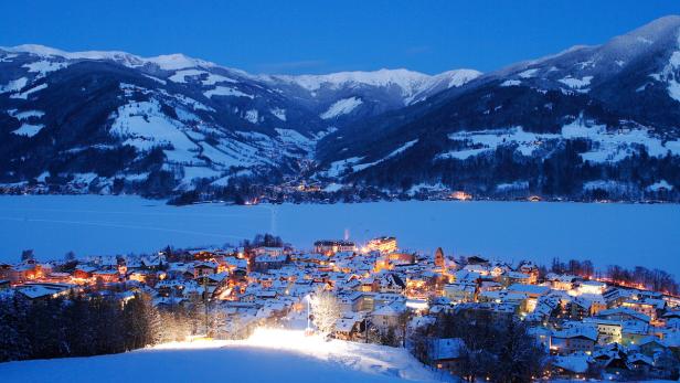 Blick auf ein verschneites Dorf am Seeufer, im Hintergrund schneebedeckte Berge.