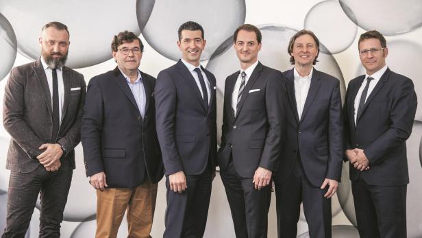 Sechs Männer in Anzügen posieren vor einer Wand mit Kreismuster.