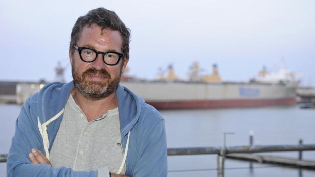 Ein Mann mit Brille und Bart steht vor einem Schiff im Hafen.