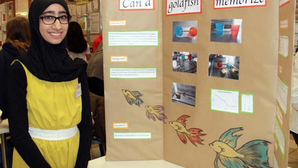 Ein junges Mädchen steht neben einer Präsentation über das Gedächtnis von Goldfischen.