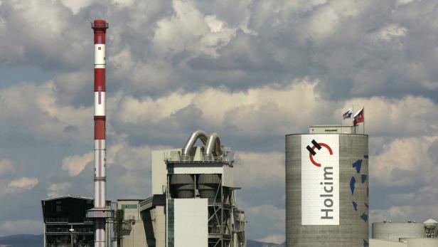 Eine Holcim Zementfabrik mit einem hohen Schornstein und einem Silo mit Schweizer Flagge vor bewölktem Himmel.