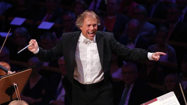 Ein Dirigent leitet ein Orchester mit einem Lächeln und ausgebreiteten Armen.