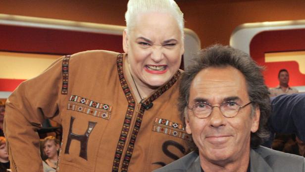 Ein Mann mit Brille lächelt, während eine Frau mit kurzem, blondem Haar grimmig dreinschaut.