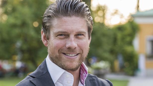 Ein lächelnder Mann mit blonden Haaren und Bart trägt ein graues Jackett.