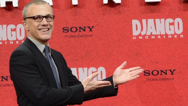 Christoph Waltz posiert vor einer Wand mit dem Logo von „Django Unchained“.