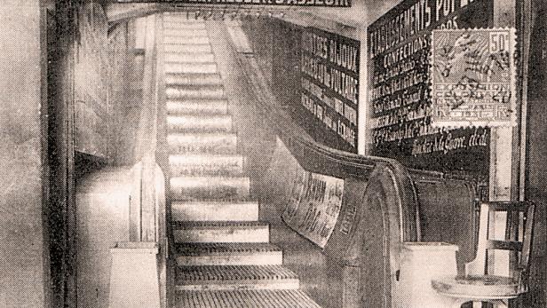 Eine alte Schwarzweißaufnahme einer Treppe mit Werbeplakaten an den Wänden.