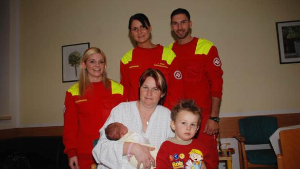 Eine Frau hält ein Neugeborenes im Arm, umgeben von zwei Sanitätern und einem kleinen Jungen.