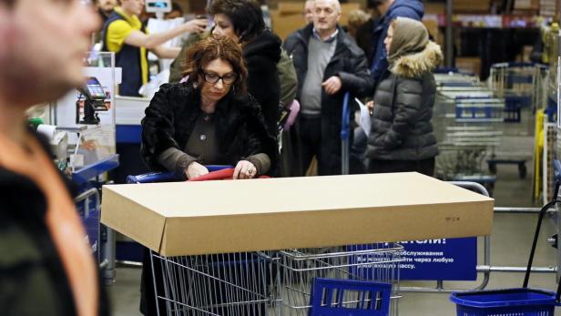 Eine Frau mit Brille schiebt einen Einkaufswagen mit einem großen Karton darin.