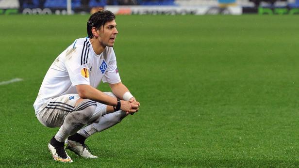 Ein Fußballspieler hockt auf dem Rasen und blickt nachdenklich zur Seite.
