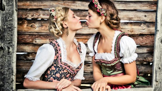 Zwei Frauen im Dirndl geben sich einen Kuss vor einer Holzwand.