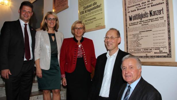 Eine Gruppe von Personen steht vor alten Plakaten, darunter Werbung für eine Kriegsanleihe und ein Wohltätigkeitskonzert.