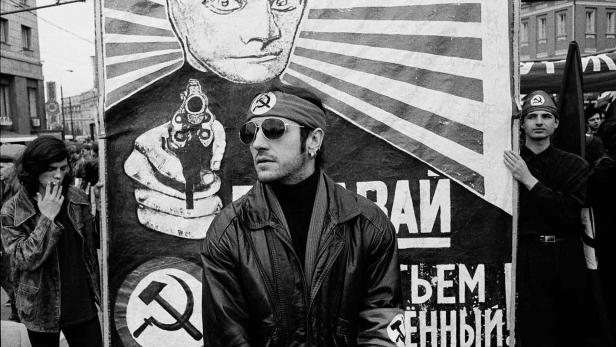 Ein Mann mit Sonnenbrille steht vor einem Plakat mit einem stilisierten Porträt und einer Pistole.