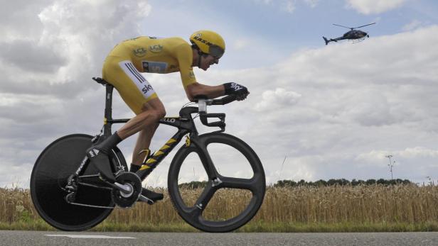 Ein Radrennfahrer in gelber Sportbekleidung fährt auf einer Landstraße, während ein Hubschrauber über ihm fliegt.