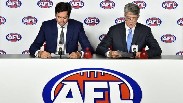 Zwei Männer in Anzügen sitzen an einem Tisch mit dem AFL-Logo und lesen Papiere.