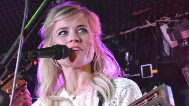 Eine blonde Frau singt mit Mikrofon und Gitarre auf einer Bühne.