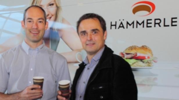Zwei Männer halten Kaffeebecher vor einem Werbeplakat von Hämmerle.