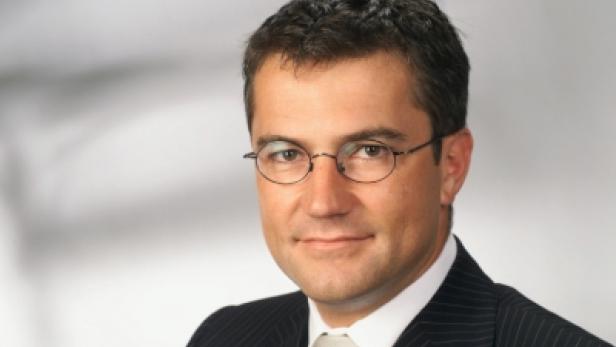 Ein Mann mit Brille und Anzug vor einem grauen Hintergrund.