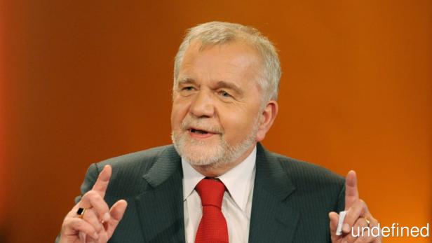 Ein Mann mit grauem Haar und roter Krawatte gestikuliert vor einem orangefarbenen Hintergrund.