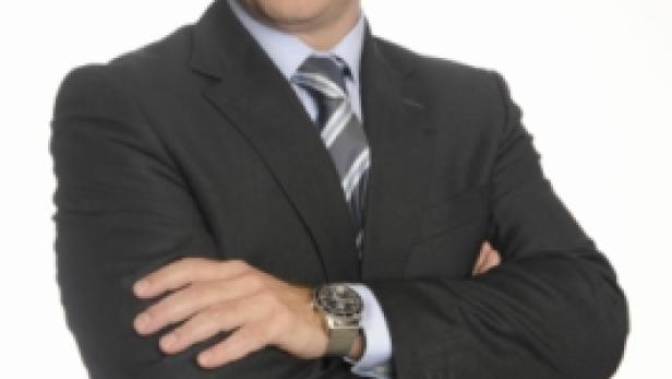 Ein Mann im Anzug mit verschränkten Armen vor einem weißen Hintergrund.
