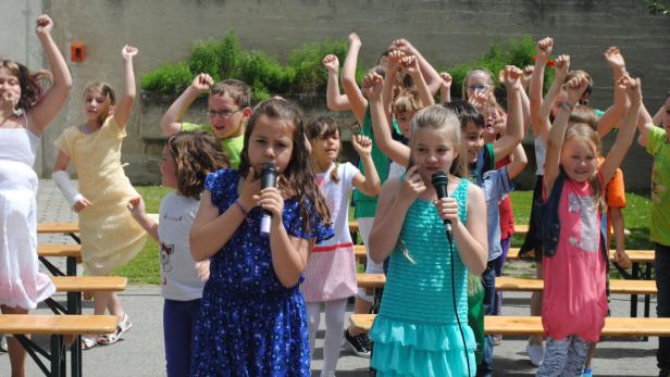 Eine Gruppe Kinder singt mit Mikrofonen und erhobenen Armen auf einer Bühne.