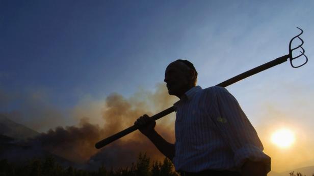 Ein Landwirt mit einer Heugabel auf der Schulter vor einem Waldbrand bei Sonnenuntergang.