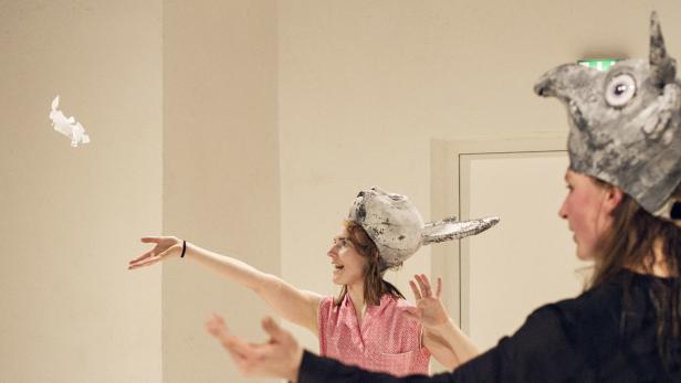 Zwei Frauen mit Vogelmasken spielen mit einem Stück Papier.