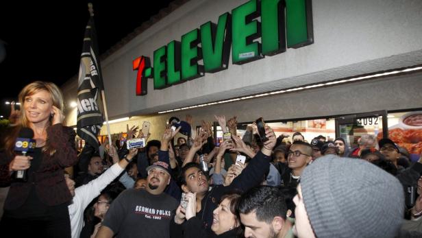 Eine Menschenmenge versammelt sich vor einem 7-Eleven-Laden, während eine Reporterin berichtet.