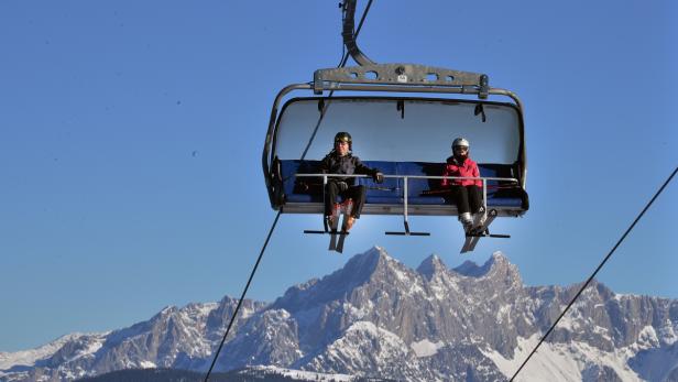Zwei Skifahrer sitzen in einem Sessellift vor einer verschneiten Bergkulisse.