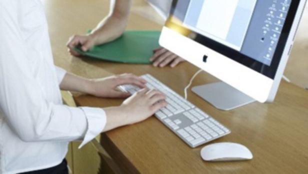 Eine Person tippt auf einer Tastatur vor einem iMac, während eine andere Person daneben steht.