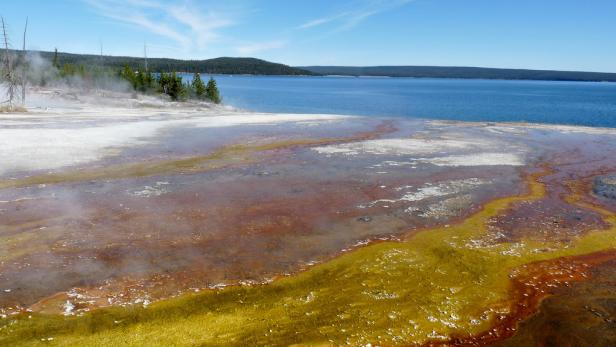 Eine farbenfrohe, dampfende Thermalquelle am Ufer eines Sees.