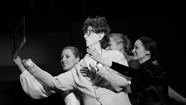 Eine Theaterszene mit einem Mann mit Brille und drei Frauen, alle lächelnd.