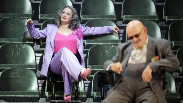 Eine Frau im lila Hosenanzug raucht auf einem Theatersessel, während ein Mann im Hintergrund sitzt.