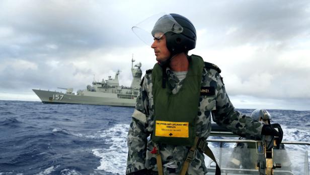 Ein Soldat in Tarnuniform und Helm auf einem Boot, im Hintergrund ein Kriegsschiff.