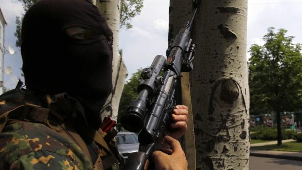 Eine vermummte Person mit einem Scharfschützengewehr steht hinter einem Baum.