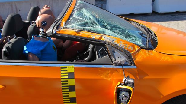 Ein orangefarbenes Auto mit Dummies und einer zerstörten Windschutzscheibe nach einem Crashtest.