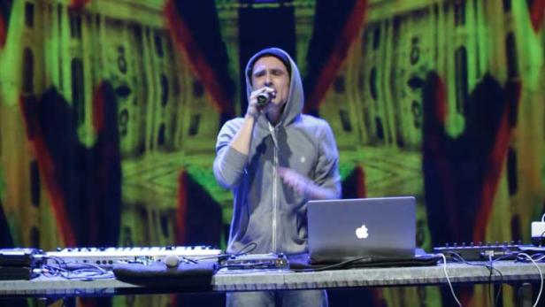 Ein Musiker mit Kapuzenpullover singt vor einem Laptop und Keyboards auf der Bühne.